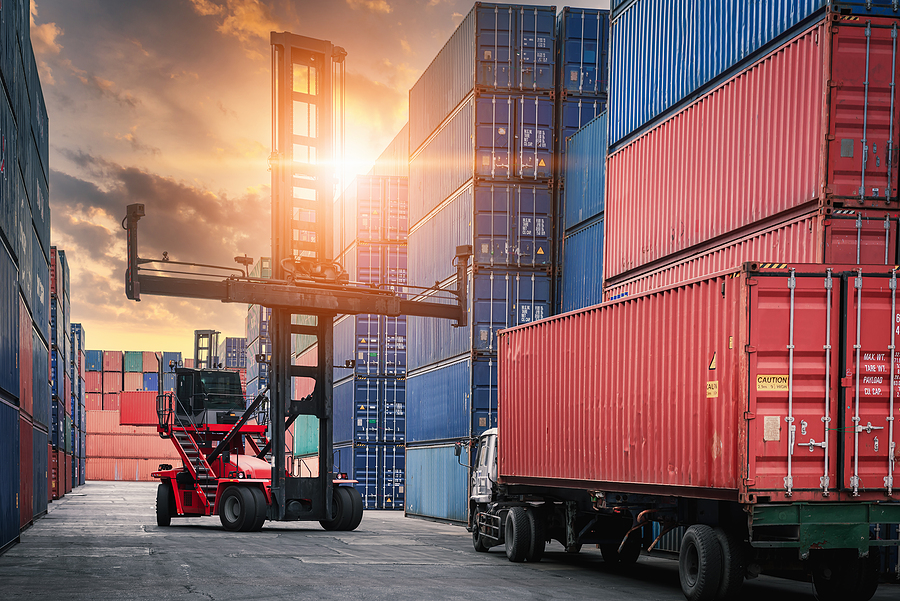 A forklift operator next to towers of shipping containers at a port at dusk