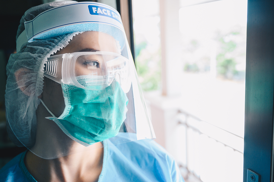 A nurse wearing a hair net, plastic goggles, a face mask, and a face shield staring exhaustedly out a window after dealing with COVID-19 patients.