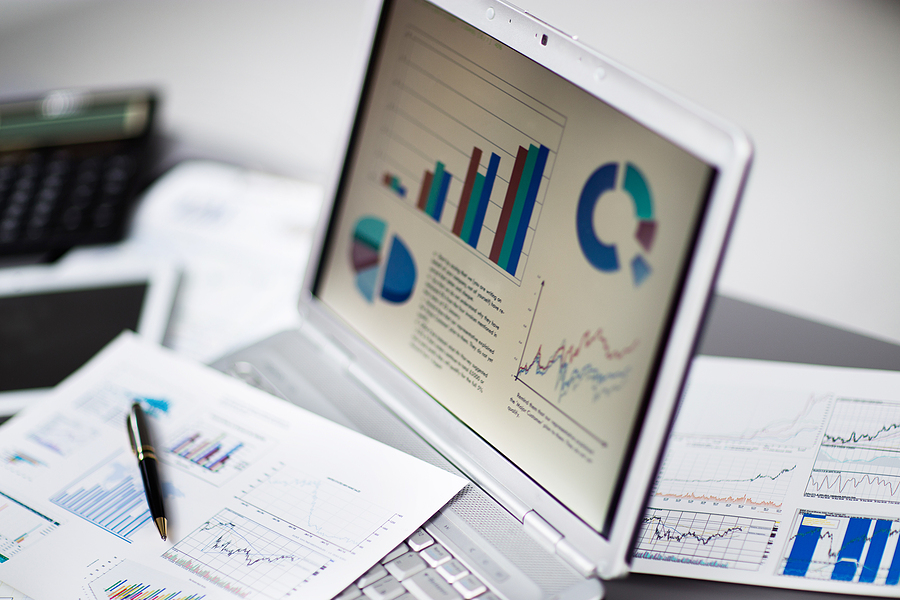 A laptop with pie and line charts on top of a desk with similar line and pie charts printed on paper strewn about.