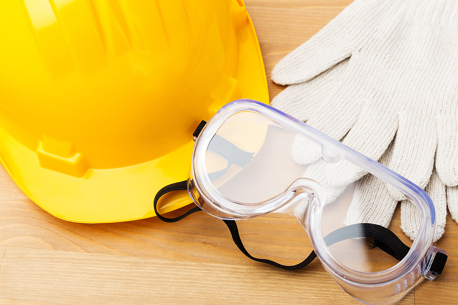 Polycarbonate safety goggles, a yellow hard hat, and white cloth gloves on top of a wooden surface.