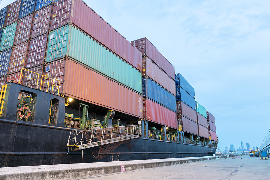 A cargo container ship at port with a city skyline in the distant background.